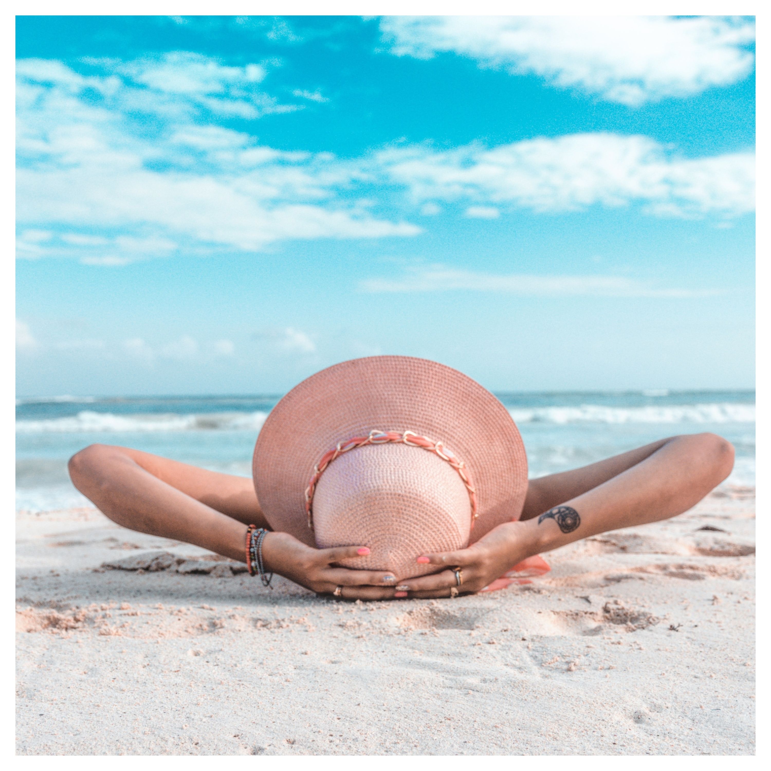 Une personne allongée sur du sable face à la mer porte un chapeau et soutient sa tête