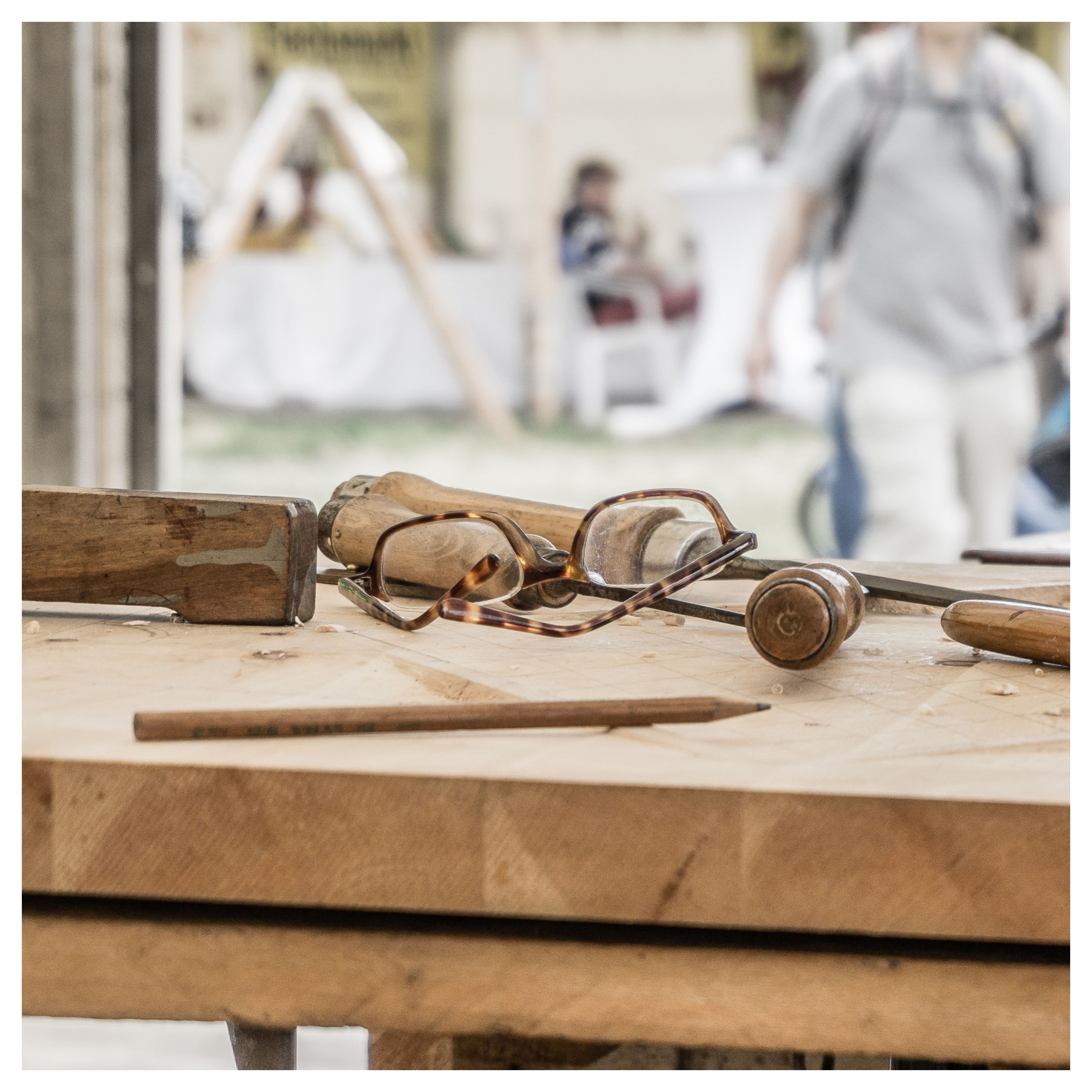Sur un établi en bois, on voit des outils (tourne-vis, crayon à papier) et une paire de lunettes de vue.