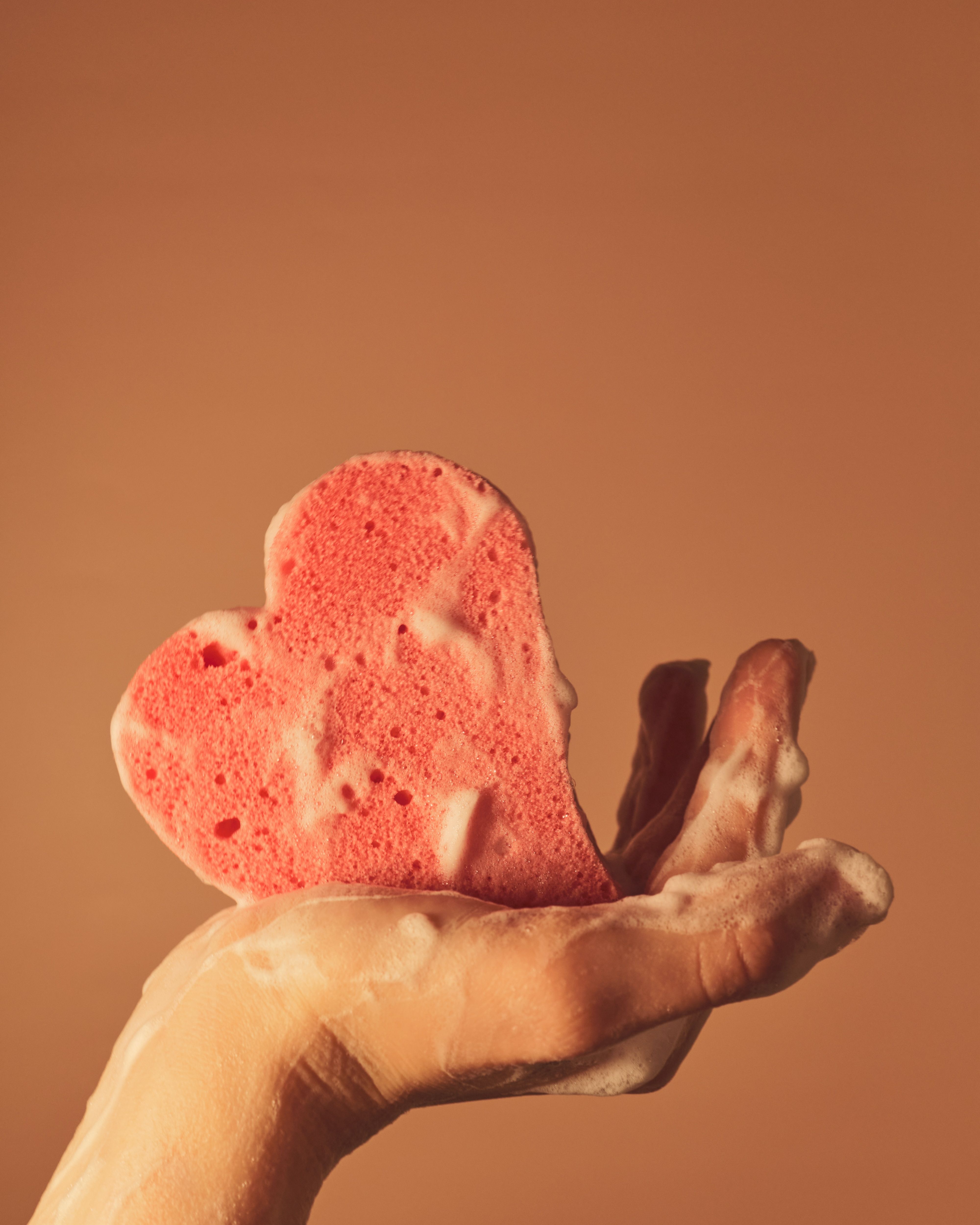 Une éponge rose en forme de coeur tenue dans une main, avec du savon