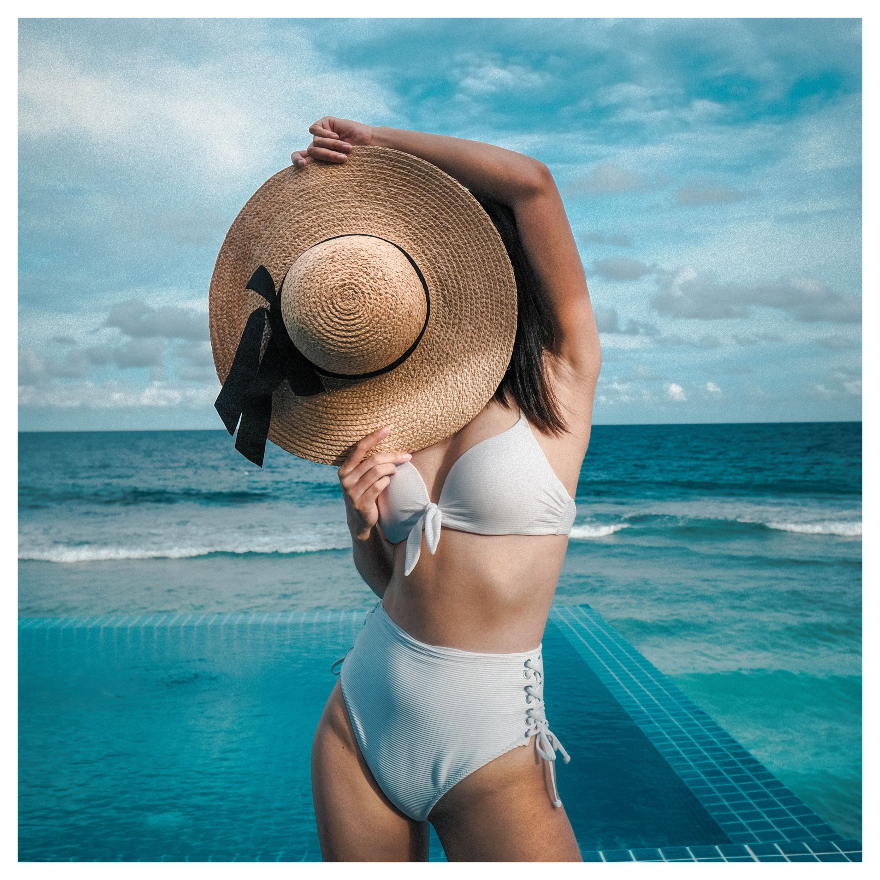 Une personne en maillot de bain blanc deux pièces qui cache son visage avec un chapeau avec la mer en arrière-plan