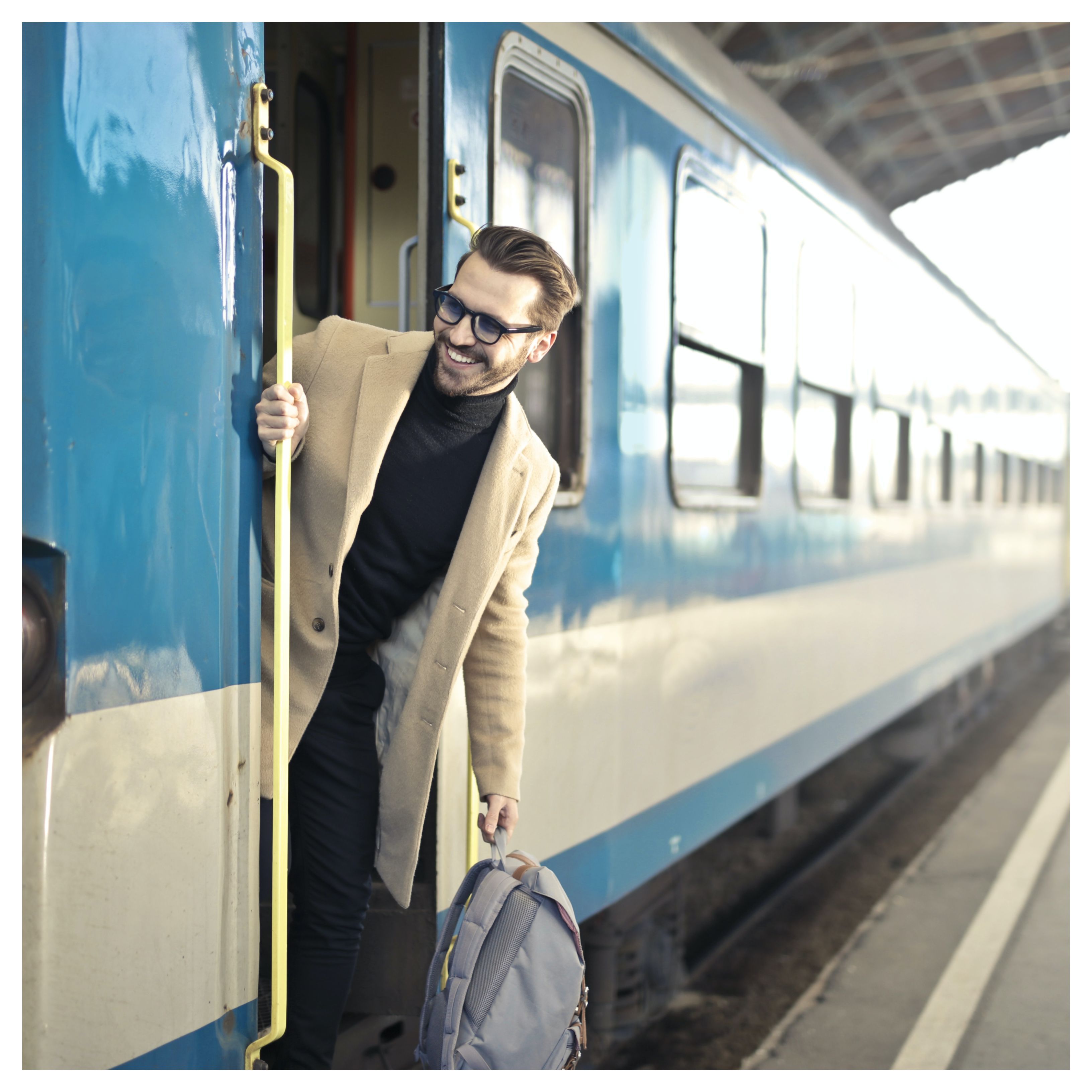 Un homme souriant se tient sur le marchepied d'un train bleu et blanc. Il tient un sac à dos gris à la main. Il porte un manteau beige et un col roulé nour. Il porte des lunettes de vue.