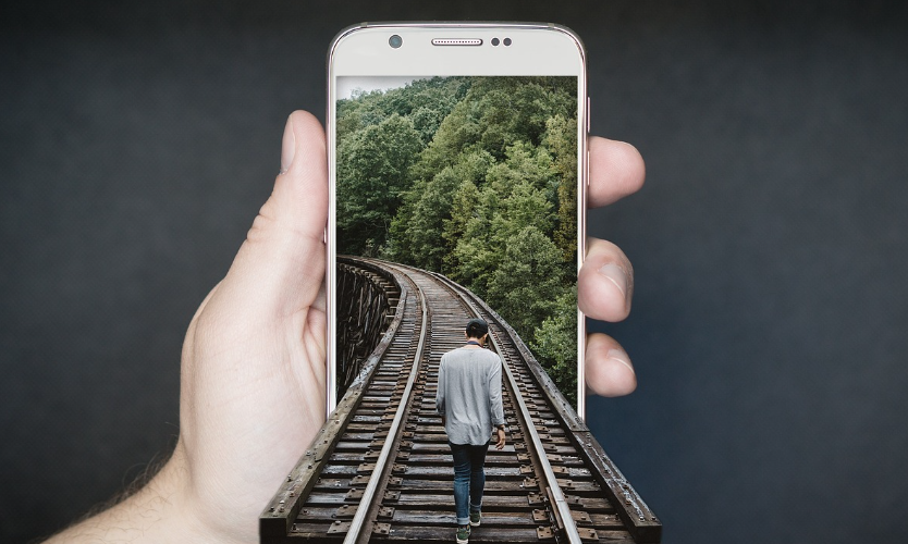 Une main tient un téléphone montrant un effet d'optique d'un homme marchant sur des chemins de fer bordé par une forêt