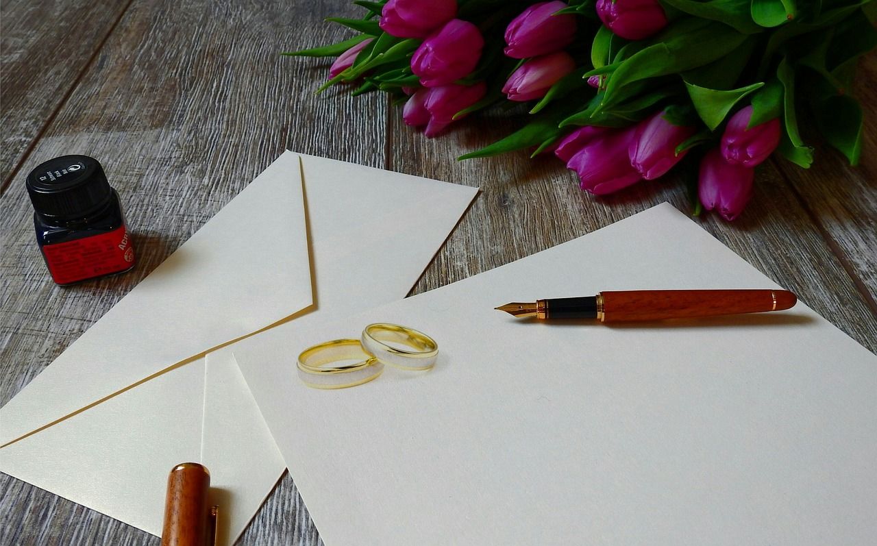 Une enveloppe blanche avec un carton blanc. Deux alliances et un stylo plume sont déposés dessus avec des fleurs roses.