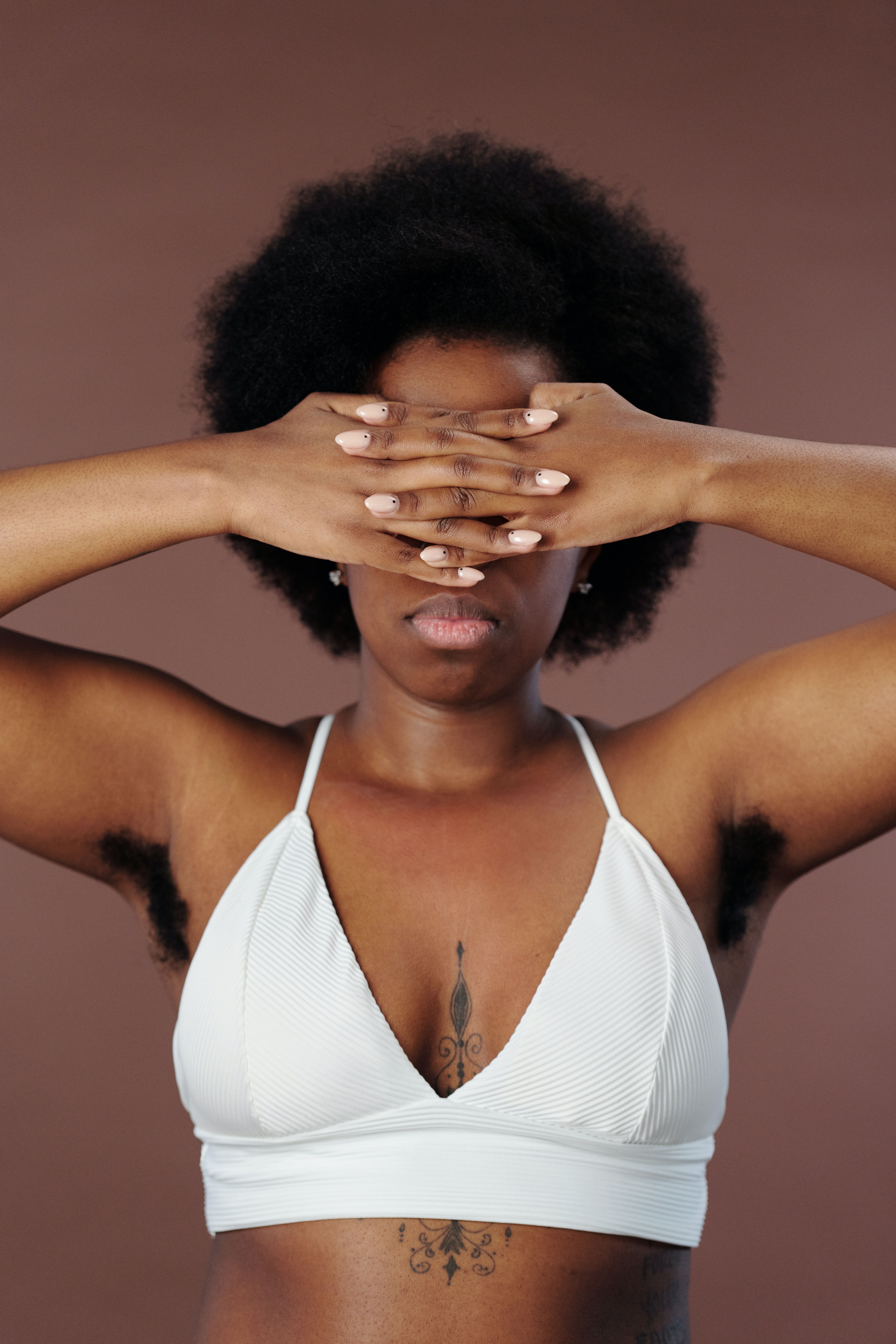Une femme noire avec un crop blanc et un tatouage inter-seins se cache les yeux avec les mains et laisse entrevoir des aisselles poilues.