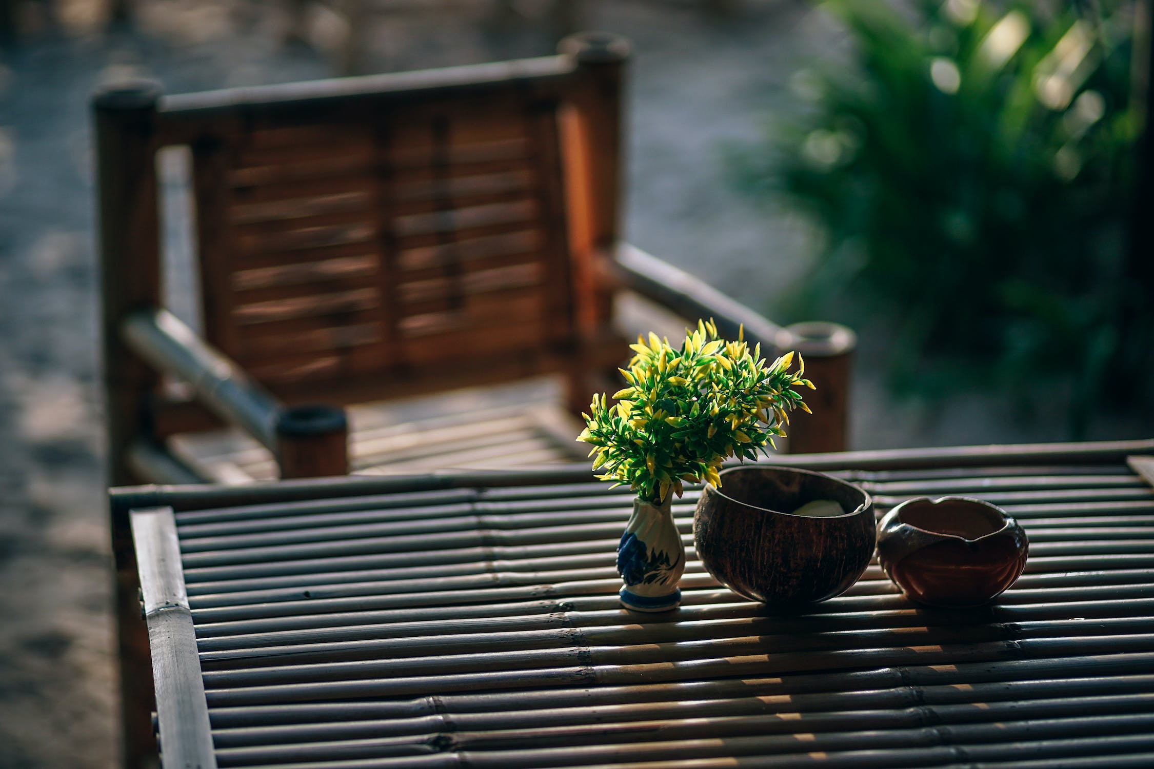 Sur l'image, on voit une table et une chaise de jardin faites en bambou. Un bouquet de feuilles de bambou est disposé dans un vase blanc, avec deux bols en bambou sur la droite.