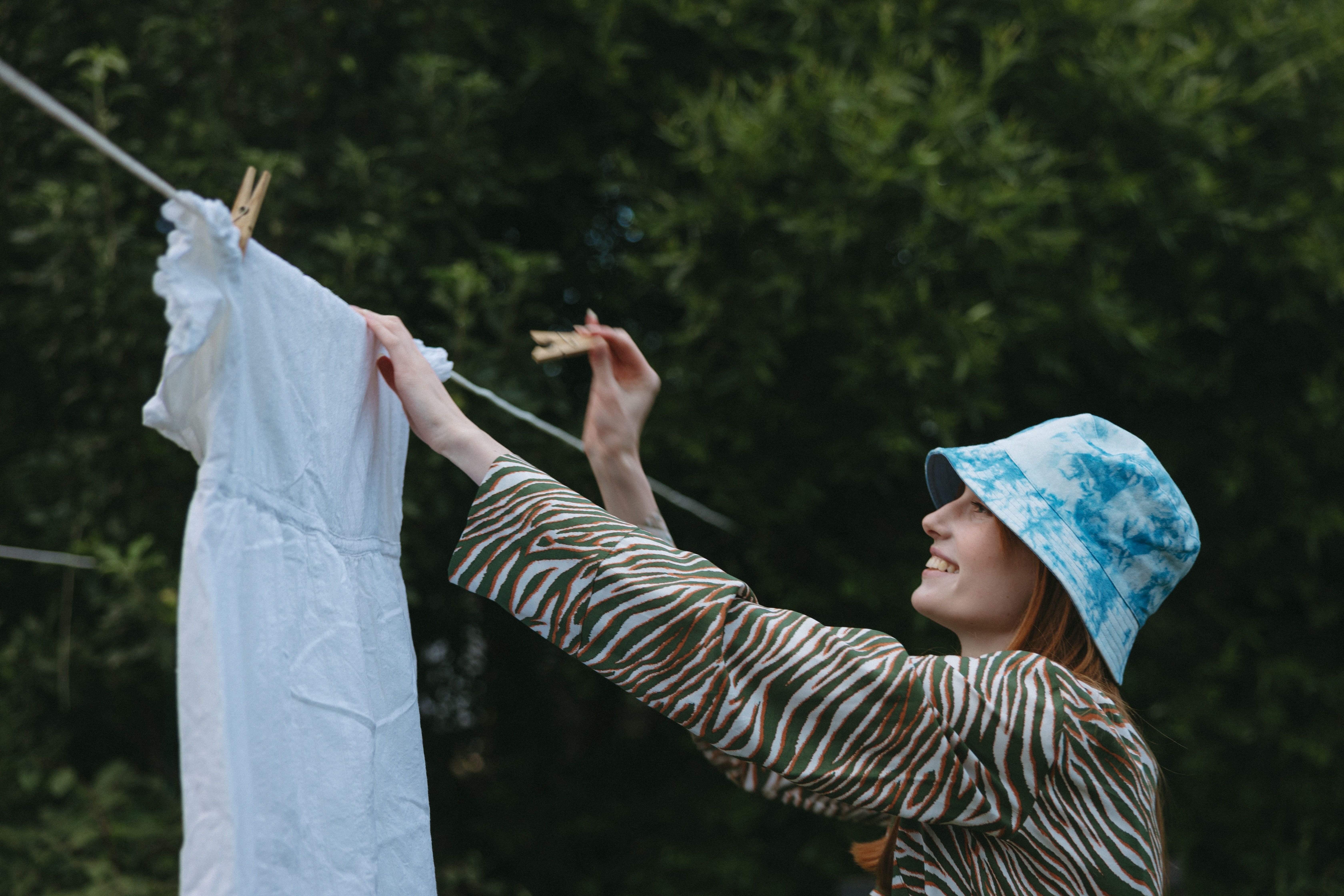 Une femme avec un bob bleu ciel délavé accroche une robe blanche pour la faire sécher, à l'aide de pinces à linge